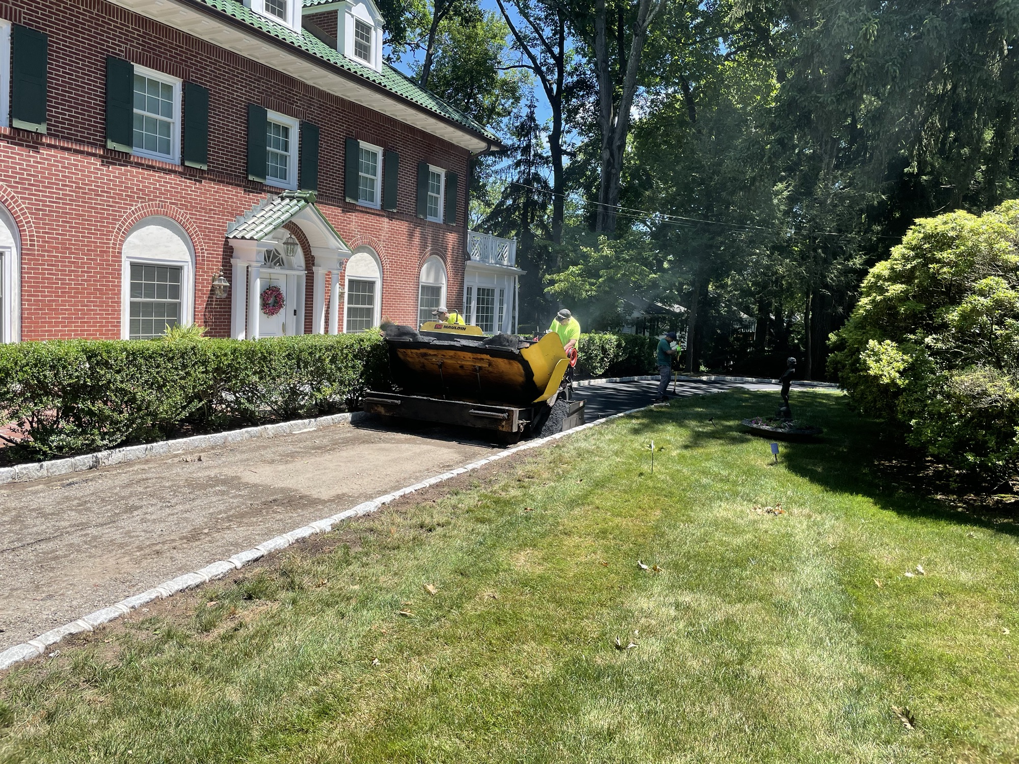 Paving crew and asphalt paving machine working on a residential driveway at a brick home